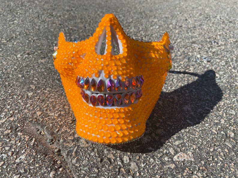 Op de afbeelding: Oranje schedelmasker bedekt met strass met een rij regenboogkleurige stenen als tanden.
