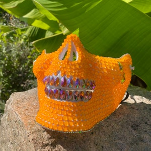Op de afbeelding: Oranje schedelmasker met witte details en iriserende strass steentjes. Het masker is gemaakt van plastic en heeft een 3D-effect.