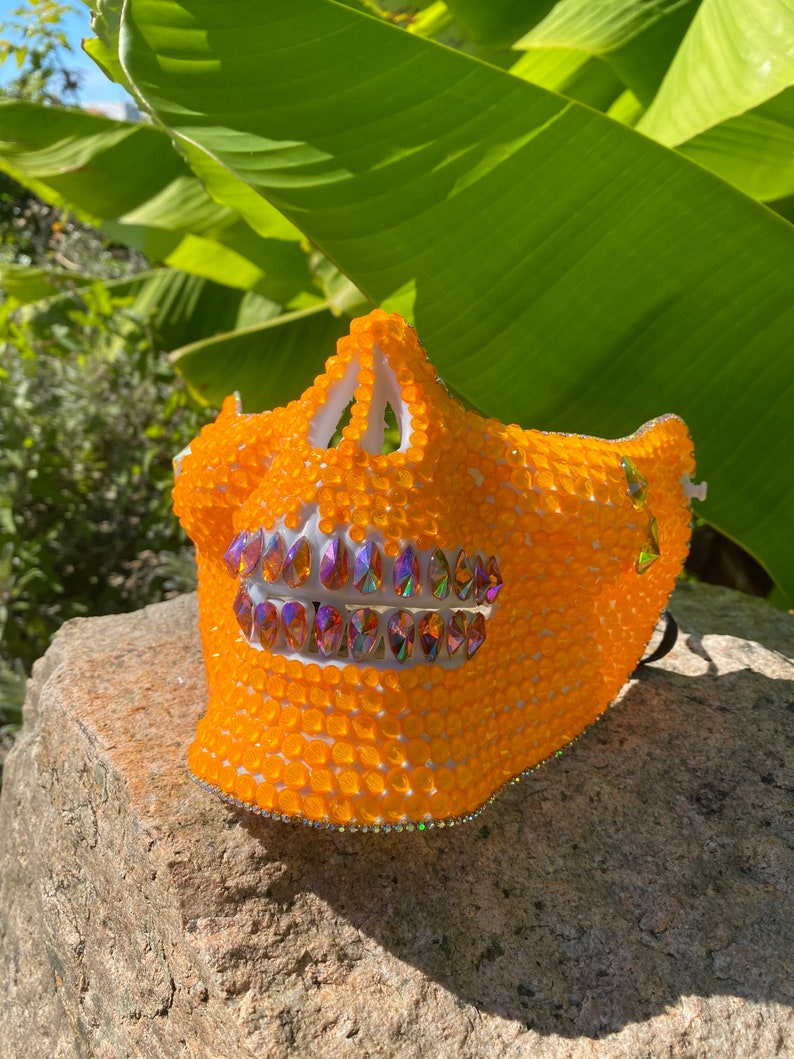 Op de afbeelding: Oranje schedelmasker met witte details en iriserende strass steentjes. Het masker is gemaakt van plastic en heeft een 3D-effect.