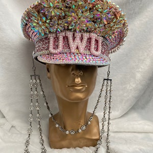 May include: A bedazzled pink and silver hat with the letters "UWU" on the front. The hat is covered in rhinestones and iridescent gems. Two strands of decorative silver chains with pearls and floral accents hang from the hat.