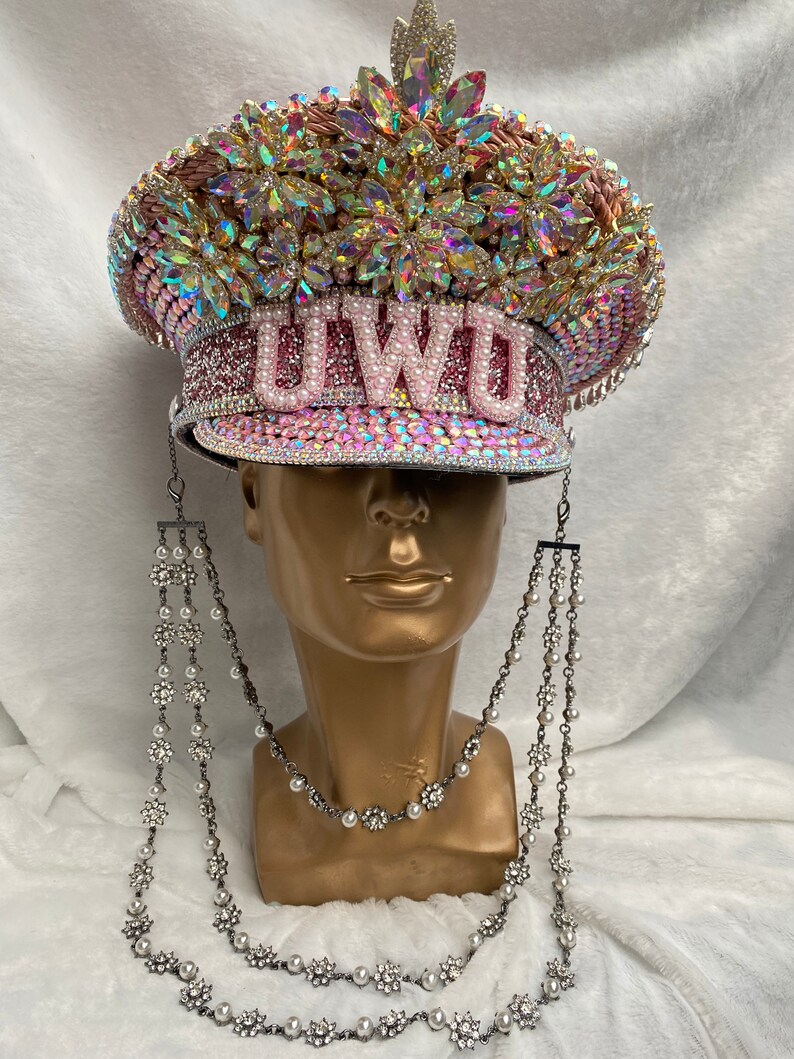May include: A bedazzled pink and silver hat with the letters "UWU" on the front. The hat is covered in rhinestones and iridescent gems. Two strands of decorative silver chains with pearls and floral accents hang from the hat.