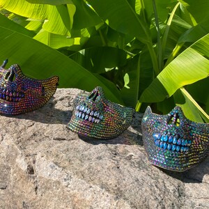 May include: Three iridescent skull masks with jewel embellishments are displayed on a gray rock. The masks have open mouths and eye sockets, and are set against a backdrop of large green leaves.