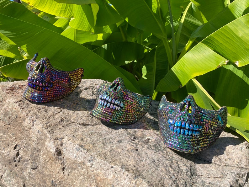 May include: Three iridescent skull masks with jewel embellishments are displayed on a gray rock. The masks have open mouths and eye sockets, and are set against a backdrop of large green leaves.