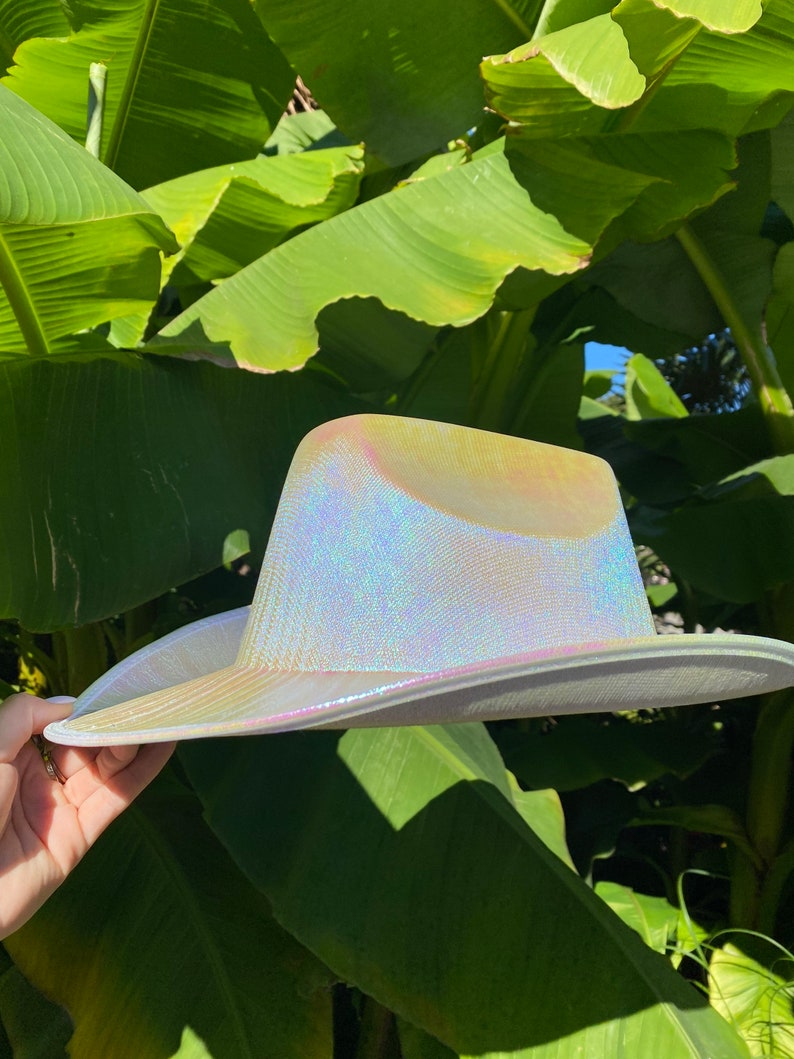 May include: A white iridescent cowboy hat with a wide brim. The hat is made of a shiny material that reflects light.