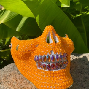 Op de afbeelding: Een wit schedelmasker bedekt met oranje en regenboogkleurige strass steentjes. Het masker heeft een gedetailleerd ontwerp en is perfect voor een kostuum of feest.