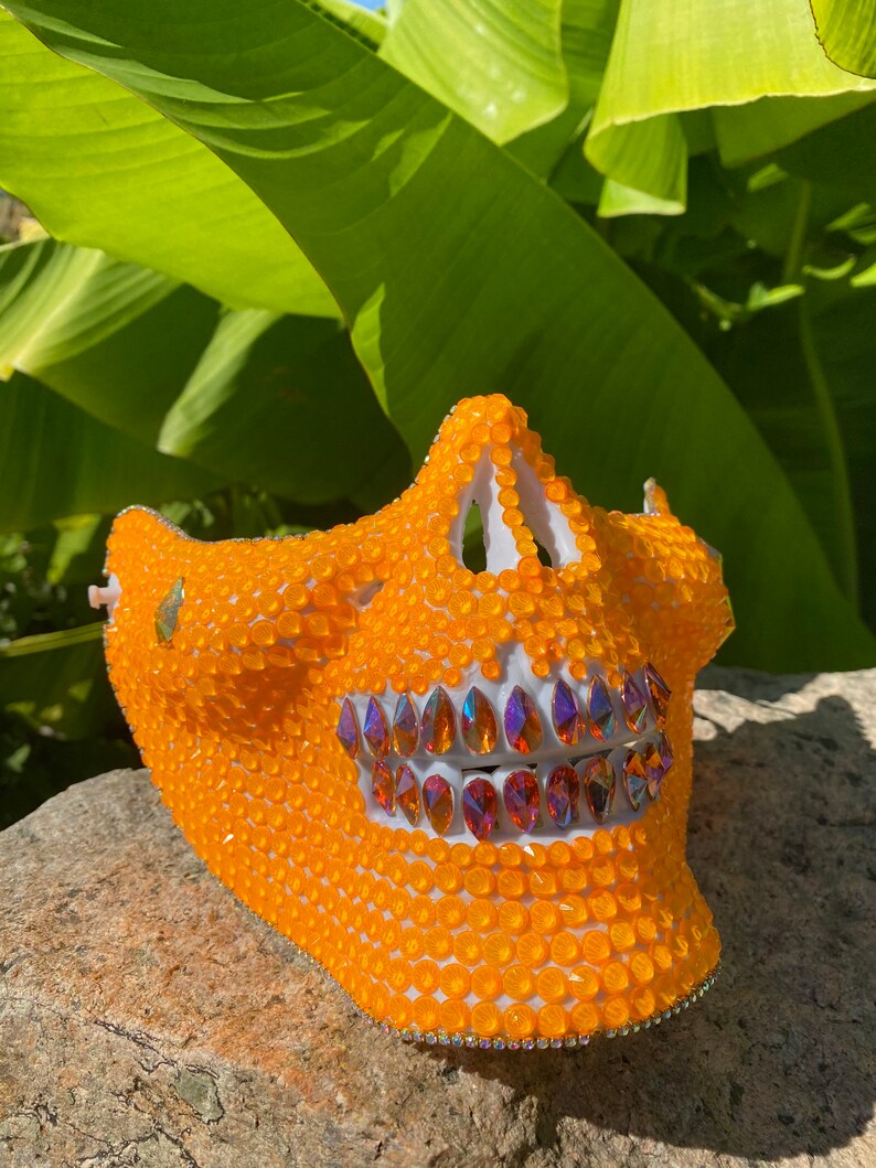 Op de afbeelding: Een wit schedelmasker bedekt met oranje en regenboogkleurige strass steentjes. Het masker heeft een gedetailleerd ontwerp en is perfect voor een kostuum of feest.