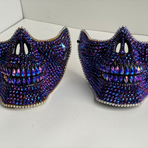May include: Two skull-shaped masks covered in iridescent blue and purple rhinestones. The masks have a black base and a white background.