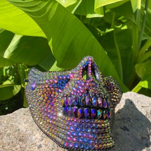 May include: A decorative skull mask covered in iridescent rhinestones. The mask features a detailed jawline and teeth, with a black trim. The rhinestones reflect a spectrum of colors, including blue, purple, and gold, creating a dazzling effect.