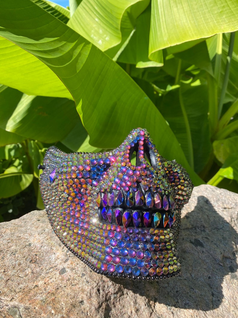 May include: A decorative skull mask covered in iridescent rhinestones. The mask features a detailed jawline and teeth, with a black trim. The rhinestones reflect a spectrum of colors, including blue, purple, and gold, creating a dazzling effect.