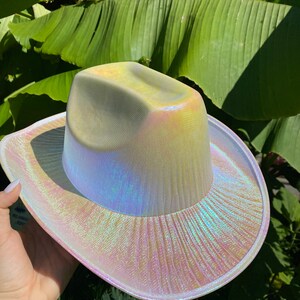 May include: A white iridescent cowboy hat with a wide brim. The hat has a subtle shimmer and a textured surface.