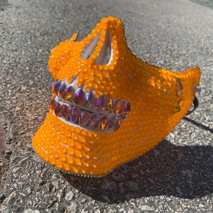 Op de afbeelding: Een feloranje schedelmasker bedekt met kleine, ronde strasssteentjes. Het masker heeft een witte basis en iriserende strasssteentjes voor de tanden.