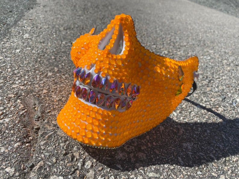 Op de afbeelding: Een feloranje schedelmasker bedekt met kleine, ronde strasssteentjes. Het masker heeft een witte basis en iriserende strasssteentjes voor de tanden.