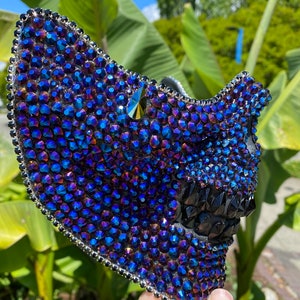 May include: A black skull mask covered in iridescent blue and purple rhinestones. The mask has a pointed top and a jagged, toothy grin.