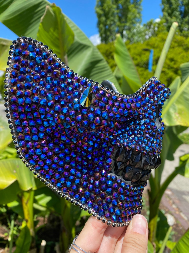 May include: A black skull mask covered in iridescent blue and purple rhinestones. The mask has a pointed top and a jagged, toothy grin.