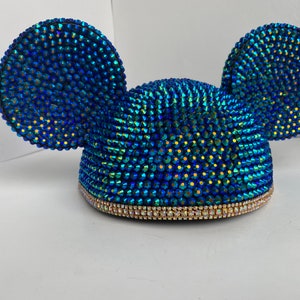 May include: A blue, bedazzled mouse ear headband. The ears and the hat are covered in iridescent blue rhinestones. The base of the hat is trimmed with a band of clear rhinestones.