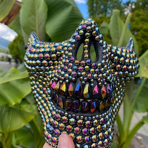 May include: A black mask covered in iridescent, multicolored beads. The mask has a skull-like design with a row of colorful, gem-like teeth. The mask is held up against a background of green foliage and a blue sky.