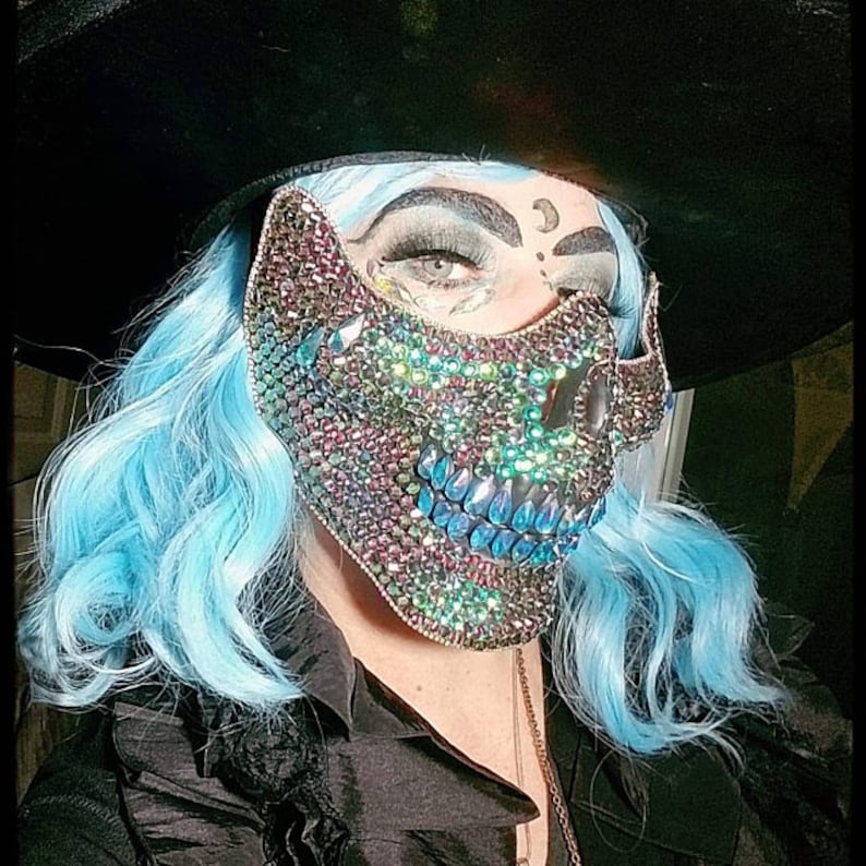 May include: A jeweled skull mask with iridescent rhinestones and blue crystal teeth. The mask is worn with a black hat and a black shirt. The person has blue hair and dramatic eye makeup. The mask is a unique accessory.