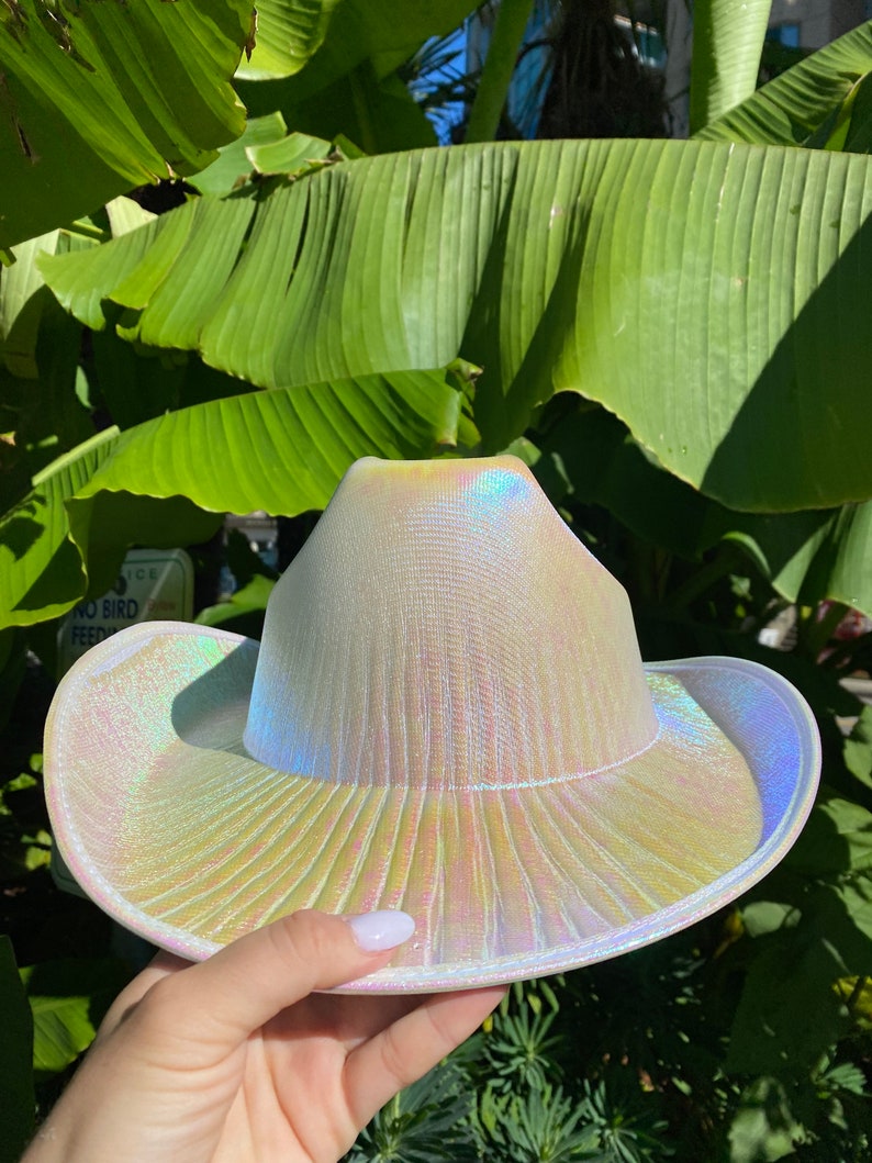 May include: A white iridescent cowboy hat with a wide brim. The hat has a textured surface and a subtle sheen.