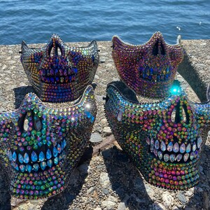 May include: Four jeweled skull masks with iridescent rhinestones. The masks are black and feature a skull design with open eye sockets and a toothy grin. The masks are displayed on a stone surface with a body of water in the background.