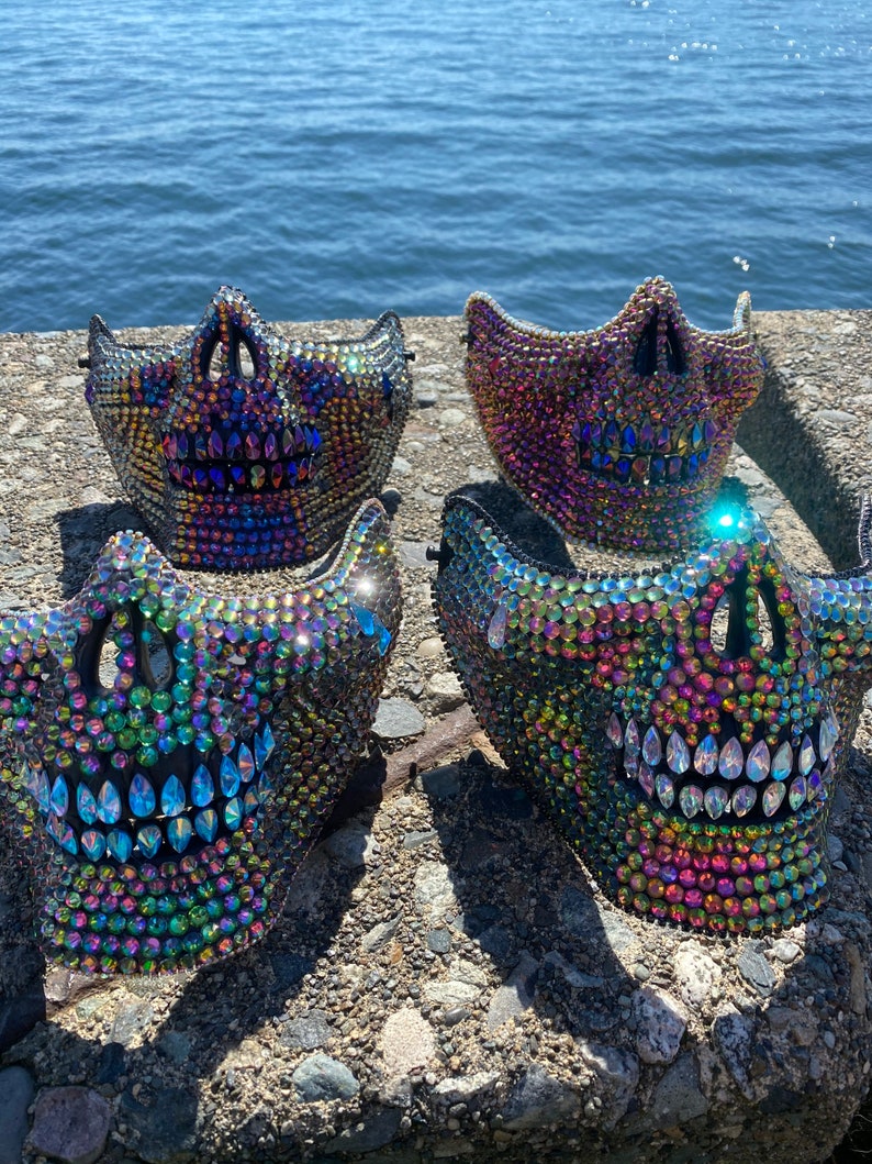 May include: Four jeweled skull masks with iridescent rhinestones. The masks are black and feature a skull design with open eye sockets and a toothy grin. The masks are displayed on a stone surface with a body of water in the background.