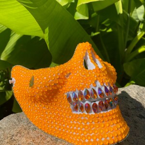 Op de afbeelding: Oranje schedelmasker bedekt met kleine oranje kralen met een rij iriserende strasssteentjes over de mond.