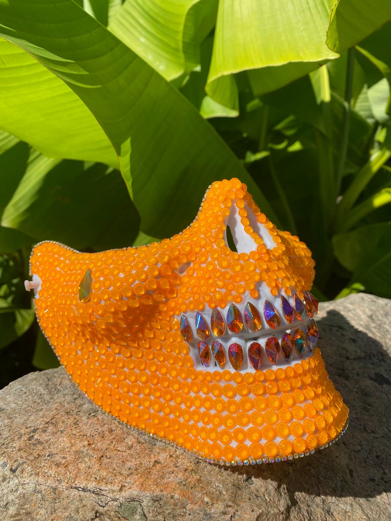 Op de afbeelding: Oranje schedelmasker bedekt met kleine oranje kralen met een rij iriserende strasssteentjes over de mond.