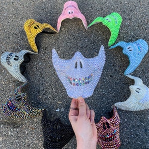 May include: A collection of bedazzled face masks in various colors, including gold, pink, green, blue, and black. The masks are decorated with rhinestones and have a skull-like design. One mask is held by a hand.
