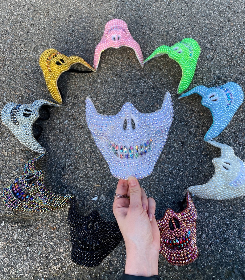 May include: A collection of bedazzled face masks in various colors, including gold, pink, green, blue, and black. The masks are decorated with rhinestones and have a skull-like design. One mask is held by a hand.