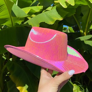 May include: A pink iridescent cowboy hat with a wide brim. The hat has a subtle shimmer and is made of a shiny material.