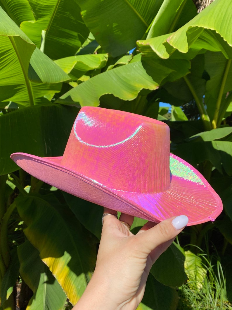 May include: A pink iridescent cowboy hat with a wide brim. The hat has a subtle shimmer and is made of a shiny material.
