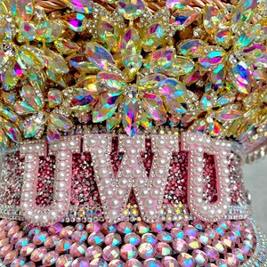 May include: A bedazzled, pink and gold decorative hat with the word "UWU" spelled out in white pearls. The hat is covered in iridescent rhinestones and crystals, with a jeweled floral design on top.