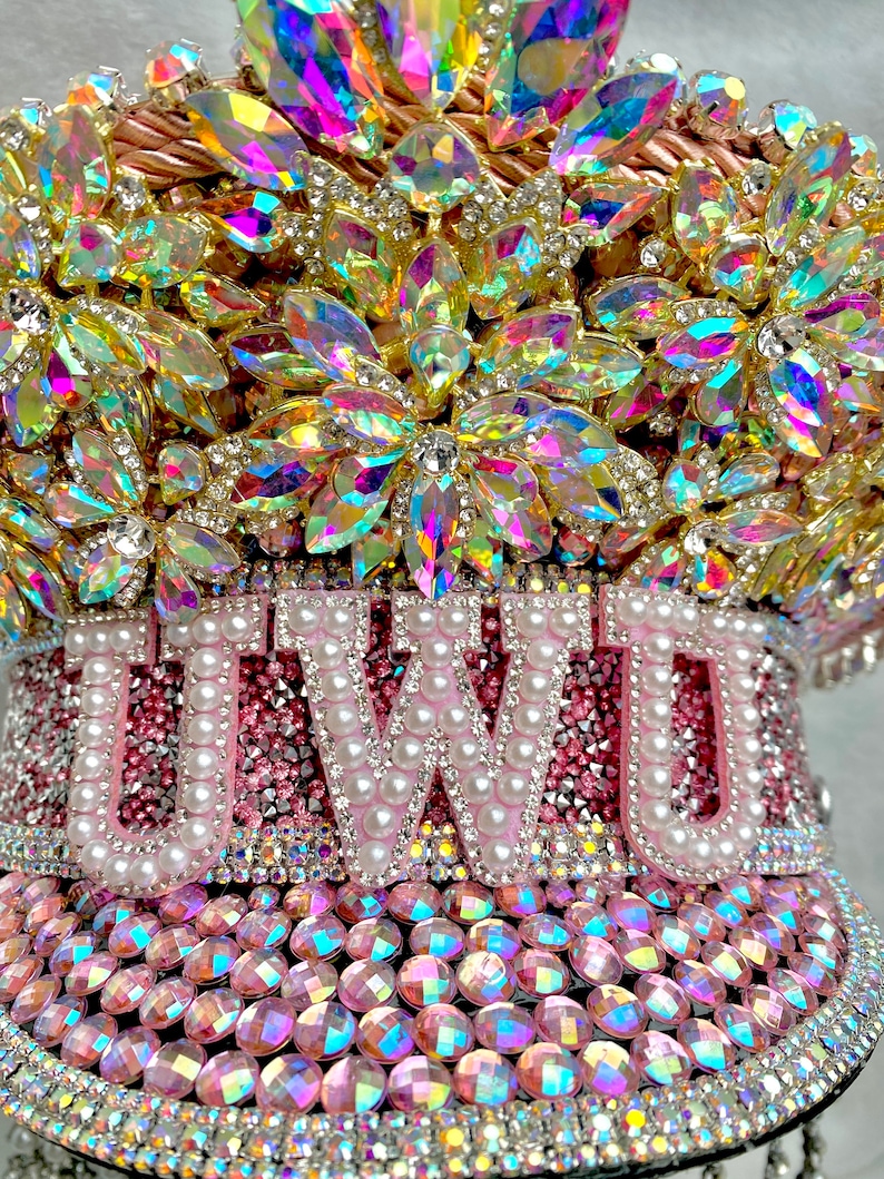 May include: A bedazzled, pink and gold decorative hat with the word "UWU" spelled out in white pearls. The hat is covered in iridescent rhinestones and crystals, with a jeweled floral design on top.