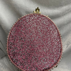 May include: A decorative, round object with a glittery pink surface, framed by a pink rope and small, silver-colored beads. A gold-colored, leaf-like embellishment sits at the top. The object is set on a brown base.