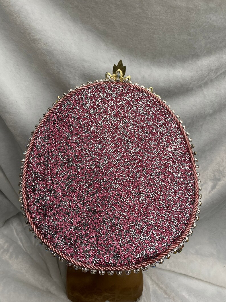 May include: A decorative, round object with a glittery pink surface, framed by a pink rope and small, silver-colored beads. A gold-colored, leaf-like embellishment sits at the top. The object is set on a brown base.
