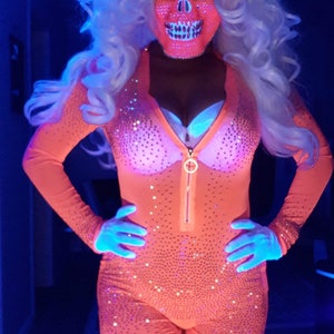 Op de afbeelding: Een persoon draagt ​​een roze en witte schedelmasker, een roze glitterpak en een wit pruik met paardenstaarten. De persoon poseert in een blauw verlichte kamer.