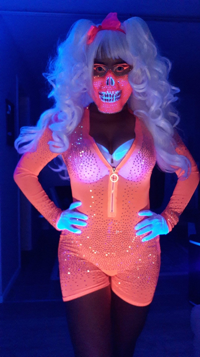 Op de afbeelding: Een persoon draagt ​​een roze en witte schedelmasker, een roze glitterpak en een wit pruik met paardenstaarten. De persoon poseert in een blauw verlichte kamer.
