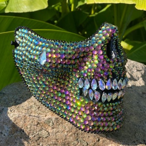 Pu&ograve; includere: Una maschera di teschio nera ricoperta di strass iridescenti. La maschera presenta un design di teschio dettagliato con una mascella e denti prominenti.