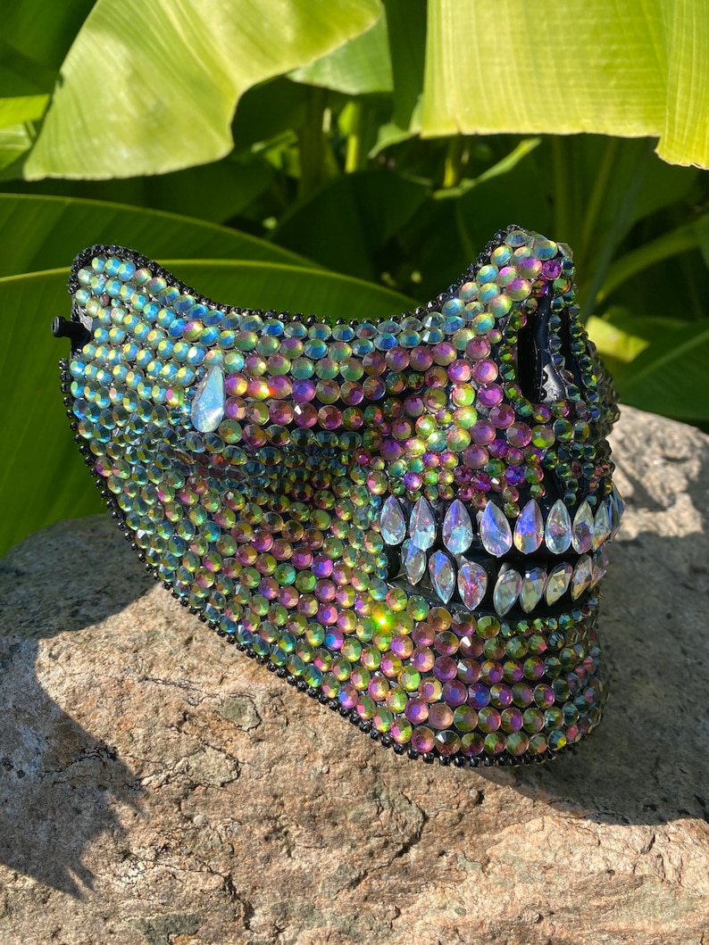 Pu&ograve; includere: Una maschera di teschio nera ricoperta di strass iridescenti. La maschera presenta un design di teschio dettagliato con una mascella e denti prominenti.