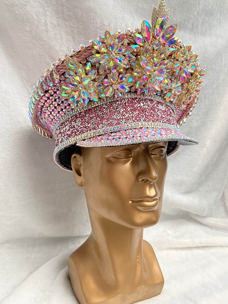 May include: A decorative pink and iridescent hat with a flat brim, heavily embellished with rhinestones and large, colorful crystals. The hat features a crown-like design on top and is displayed on a gold mannequin head.
