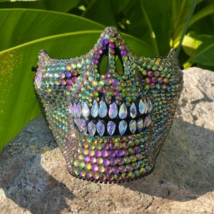 Pu&ograve; includere: Una maschera di teschio nera ricoperta di strass verdi, viola e trasparenti iridescenti.