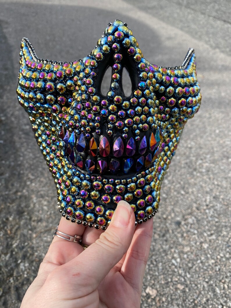 May include: A black, half-face mask, heavily embellished with iridescent, multicolored rhinestones. The mask features a skull-like design with a prominent jawline and pointed teeth. The mask is held by a hand.
