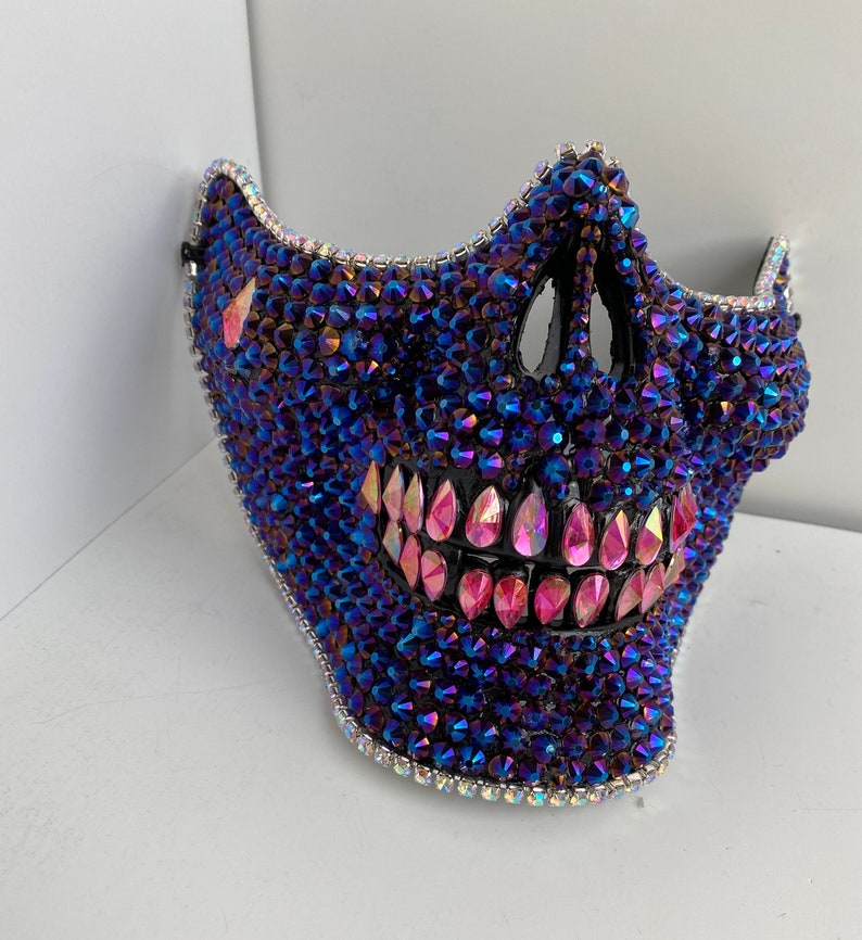 May include: A skull-shaped mask covered in iridescent blue and pink rhinestones. The mask has a wide, toothy grin.