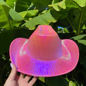 May include: A pink iridescent cowboy hat with a wide brim. The hat has a shiny, holographic finish.