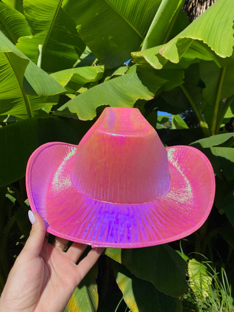May include: A pink iridescent cowboy hat with a wide brim. The hat has a shiny, holographic finish.