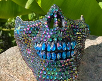 May include: A black skull mask covered in iridescent rhinestones with a row of blue teardrop-shaped rhinestones across the mouth.