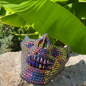 May include: A decorative skull mask covered in iridescent rhinestones. The mask features a detailed design with a prominent jawline and eye sockets. The rhinestones reflect a spectrum of colors, including purple, blue, and gold, creating a dazzling effect.