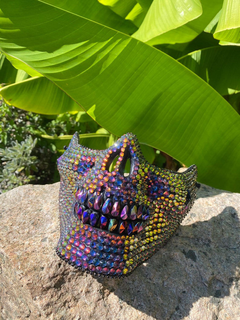 May include: A decorative skull mask covered in iridescent rhinestones. The mask features a detailed design with a prominent jawline and eye sockets. The rhinestones reflect a spectrum of colors, including purple, blue, and gold, creating a dazzling effect.