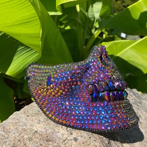 May include: A decorative mask covered in iridescent rhinestones. The mask is primarily purple, blue, and gold, with a black border. The mask is resting on a gray stone, with green leaves in the background.