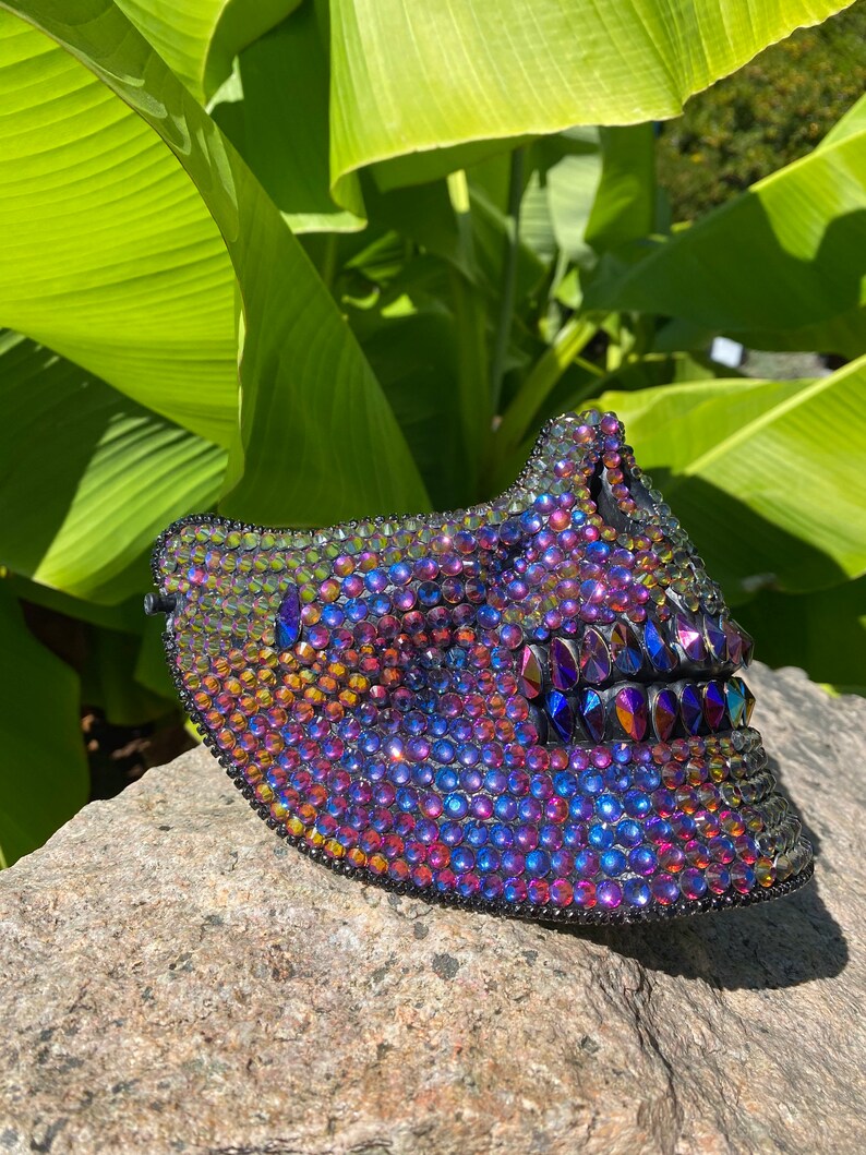 May include: A decorative mask covered in iridescent rhinestones. The mask is primarily purple, blue, and gold, with a black border. The mask is resting on a gray stone, with green leaves in the background.