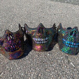Pu&ograve; includere: Tre maschere di teschio iridescenti con strass. Le maschere sono nere con un arcobaleno di colori negli strass. Le maschere sorridono tutte.